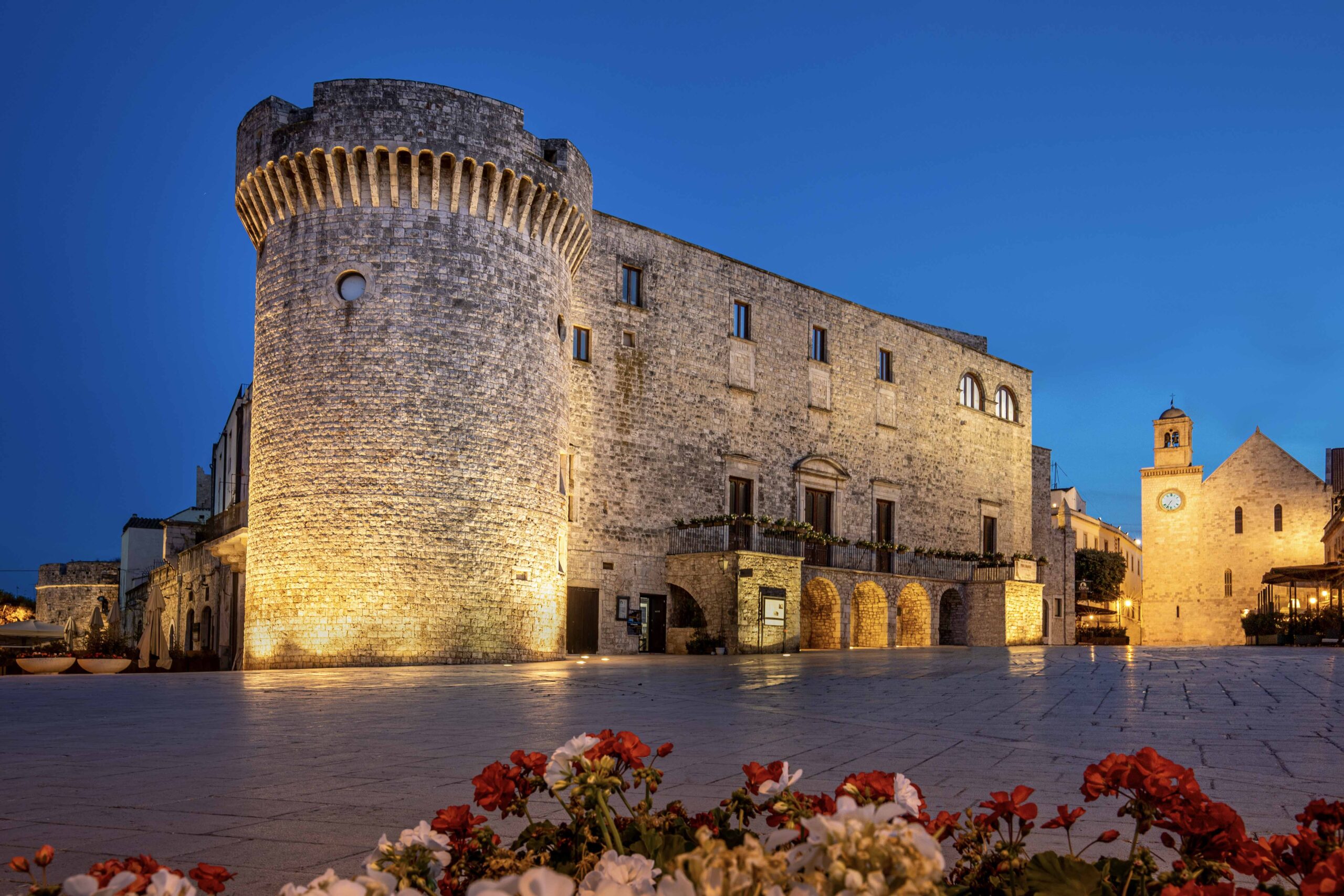 Veduta serale del Castello di Conversano illuminato, con la piazza antistante e la Cattedrale sullo sfondo, incorniciata da fiori in primo piano.