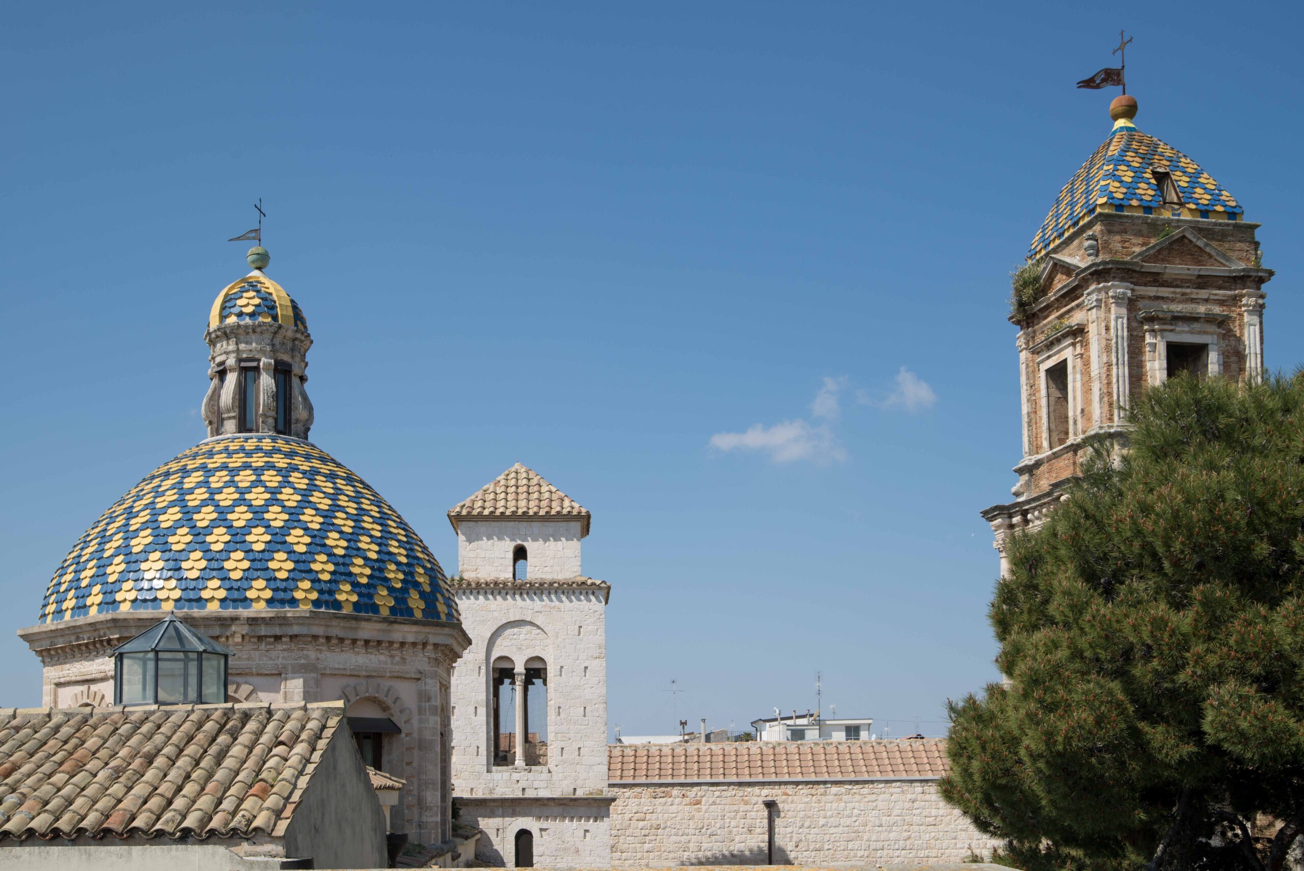 La colorata cupola e il campanile storici di Conversano ripresi in una luminosa giornata estiva, con dettagli architettonici in pietra e mattoni sotto un cielo azzurro.